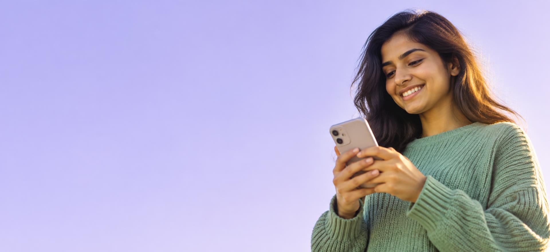 A young woman smiling at her phone against a clear lavender sky